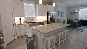 Uma cozinha moderna com armários brancos, uma ilha de granito com banquetas, eletrodomésticos de aço inoxidável e iluminação pendente.