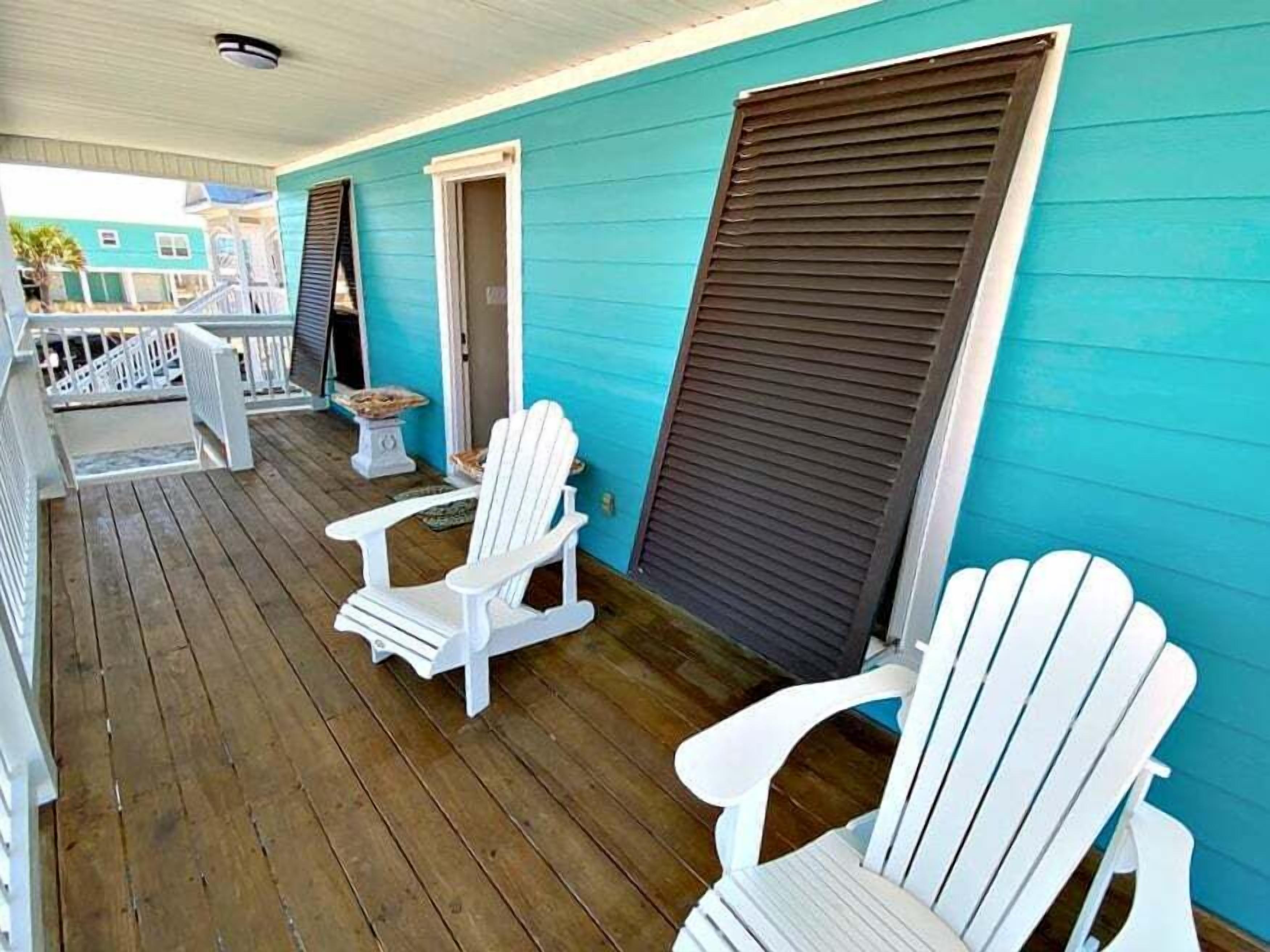 Eine überdachte Veranda mit weißen Adirondack-Stühlen, einem kleinen Tisch und Blick auf ein blaues Gebäude im Hintergrund.
