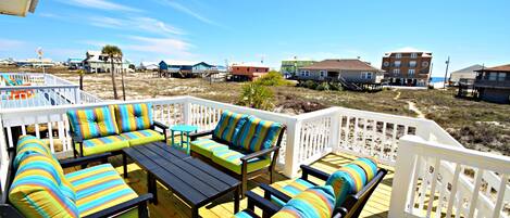 A wooden deck with outdoor furniture, including striped sofas and a table, overlooking a beach area with buildings and a clear sky.