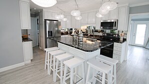 Une cuisine moderne avec des meubles blancs, un grand îlot central doté de tabourets, des appareils en acier inoxydable et des lustres en cristal.