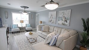 Un salon avec un canapé beige, un tapis à motifs et deux fauteuils. Il y a deux tableaux au mur, un ventilateur de plafond et une fenêtre avec des stores.