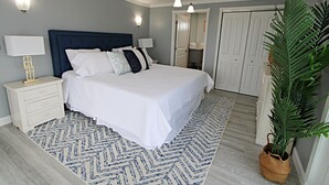 Une chambre à coucher avec un lit, des tables de chevet, une grande plante en pot et un tapis à motifs.