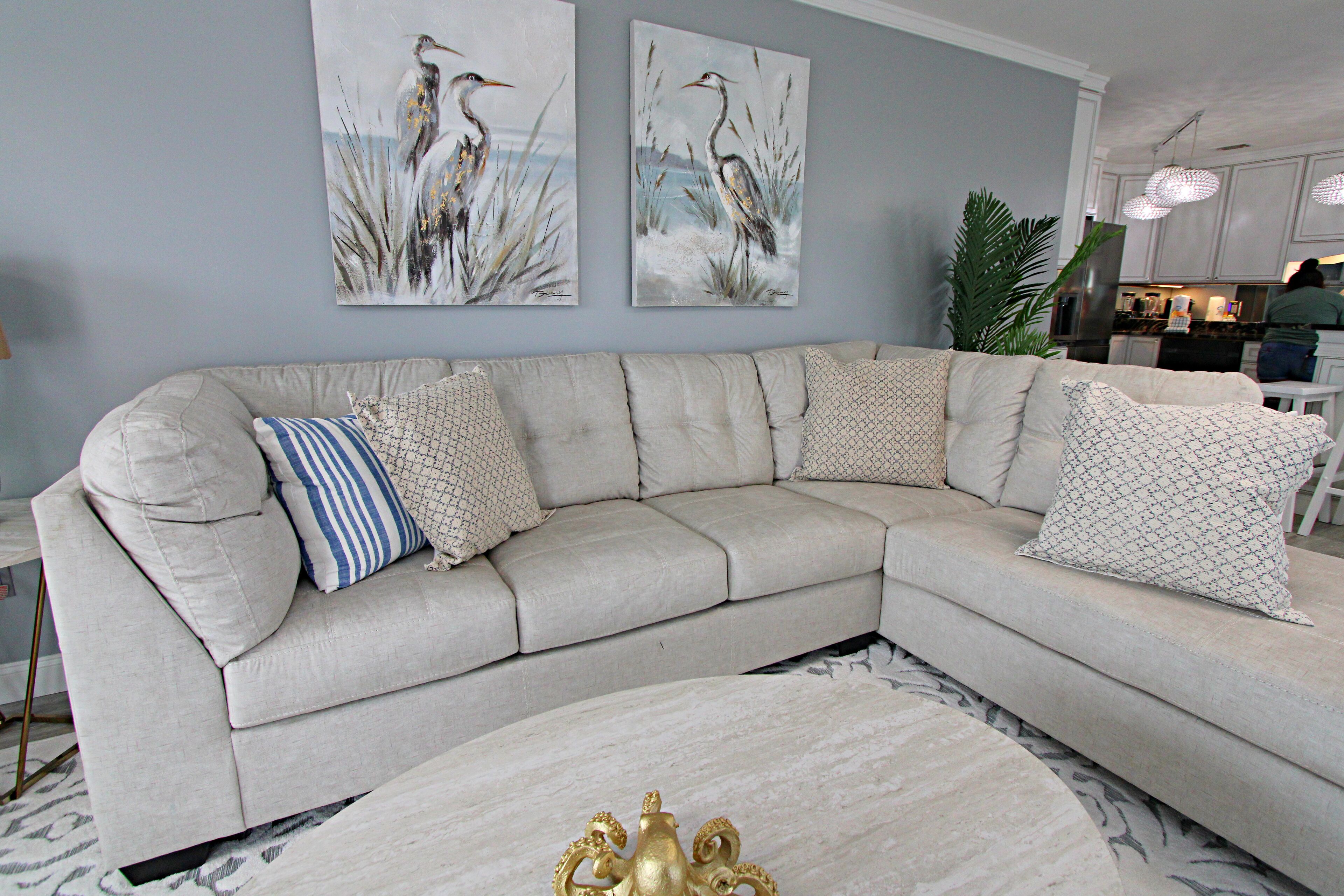 A living room with a beige sectional sofa, a round coffee table with a gold decorative piece, and two wall-mounted paintings of birds.