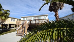 Une propriété située en bord de mer, dotée d’un escalier en bois menant à un balcon, entourée de palmiers et offrant une vue sur un ciel dégagé.