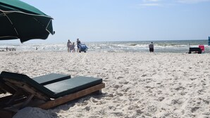 Ein Strand mit einer Liege, einem Sonnenschirm, einer Person, die spazieren geht, und einem Jetski im Hintergrund.