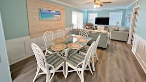 A dining area with a glass table and white chairs, a ceiling fan, and a TV in the background. - Stay Salty (Gulf Shores)