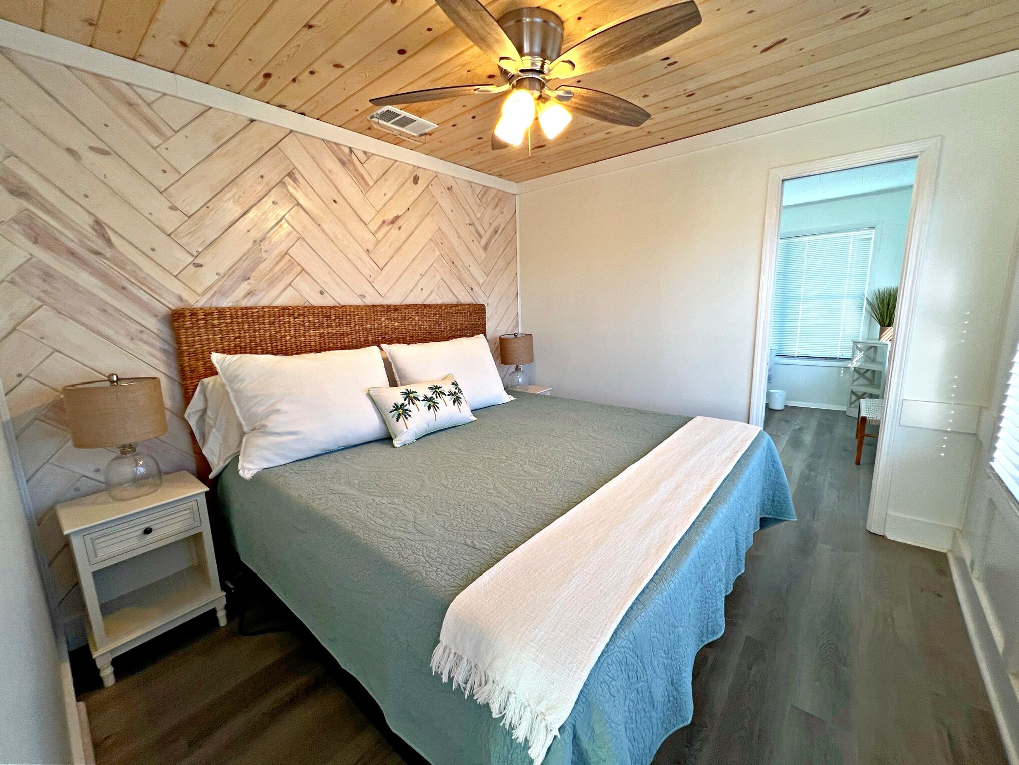 A bedroom with a herringbone-patterned ceiling, a bed with a woven headboard, white bedding, and a ceiling fan.