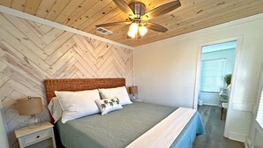 A bedroom with a herringbone-patterned ceiling, a bed with a woven headboard, white bedding, and a ceiling fan. - Stay Salty (Gulf Shores)