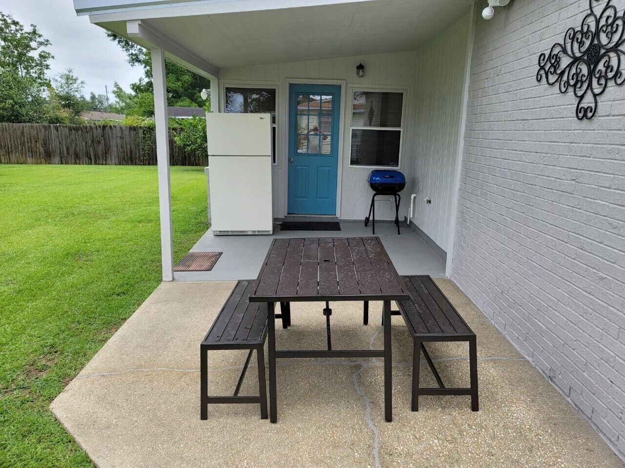 Un patio al aire libre con una mesa y bancos de madera, una puerta azul y una parrilla para barbacoa.