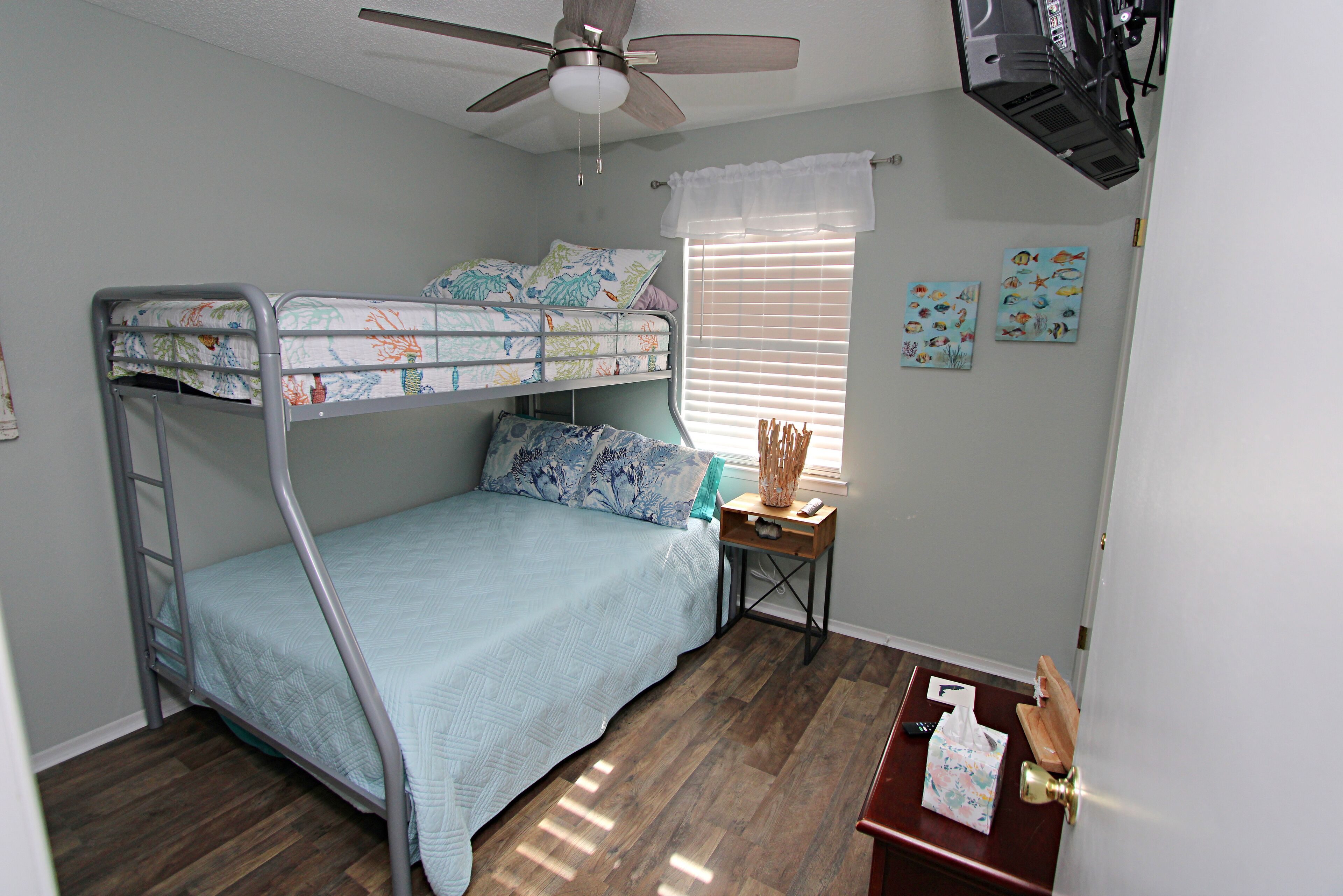 A bunk bed room with a ceiling fan, a small table with a lamp, and artwork on the wall.