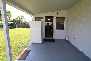 Un patio couvert équipé d’un réfrigérateur, d’une porte donnant sur un espace intérieur et offrant une vue sur une cour verdoyante.