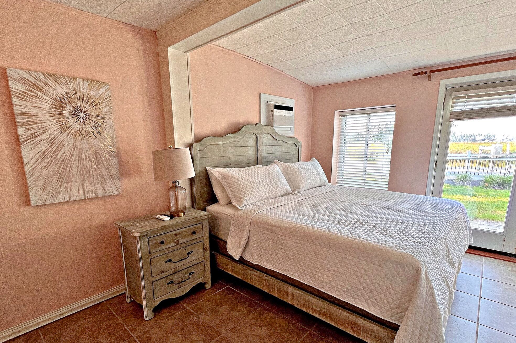 A bedroom with a bed, a nightstand, a lamp, a wall clock, a window with blinds, and a view of the outdoors.