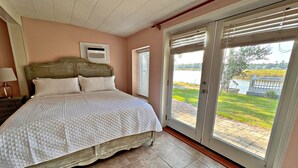 A bedroom with a bed, a nightstand, a window with blinds, and a view of a lake. - Porpoise Pass - Ground Floor Guest Suite (Foley)