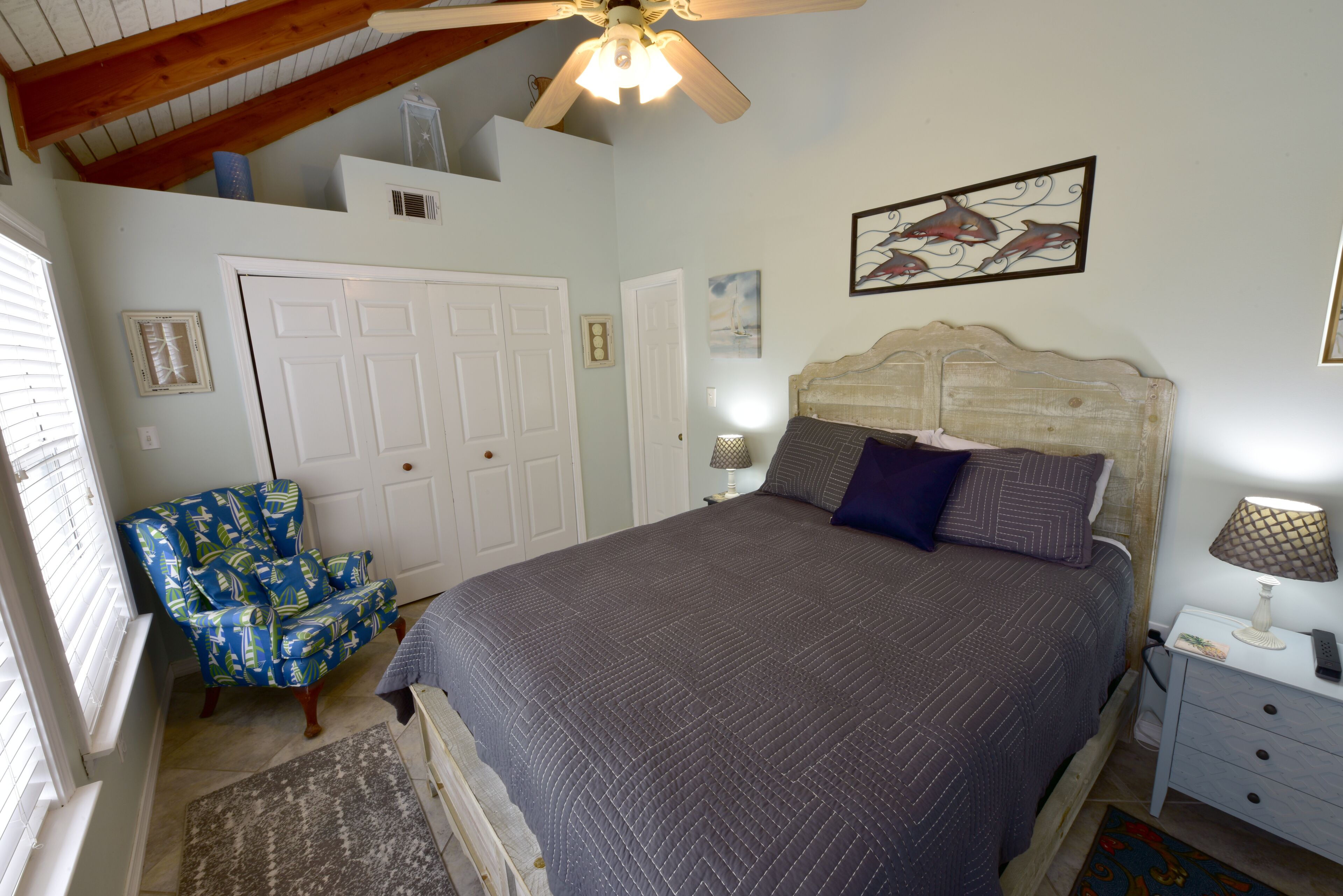 A bedroom with a large bed, a wooden headboard, a ceiling fan, a chair, a nightstand, and a framed picture on the wall.