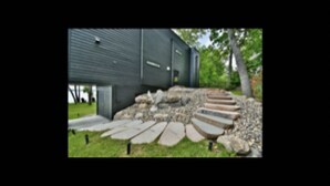 A building with a dark exterior, a stone pathway leading to a door, and a tree in the background. - Spectacular Waterfront Home  (Orono)