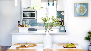 Een keuken met een wit aanrecht, een witte vaas met groene planten en een houten snijplank met etenswaren.