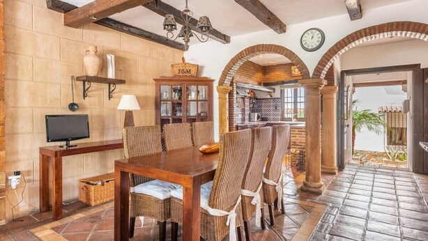 Una zona de menjador amb taula de fusta i cadires de vimetla, televisió i una cistella penjada a la paret amb la inscripció "F&M".