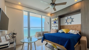 Ein modernes Schlafzimmer mit einem großen Bett, einem Deckenventilator, Blick auf den Ozean, einem kleinen runden Tisch und einem Fernseher.