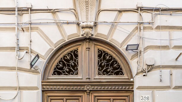 Una puerta de madera grande y ornamentada con un arco decorativo encima, en un edificio con fachada texturizada.