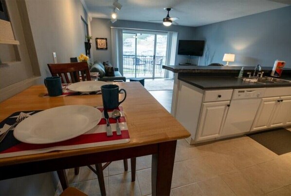 A dining area with a wooden table set for two, a kitchen with white cabinets, and a view of the outdoors through a sliding glass door.