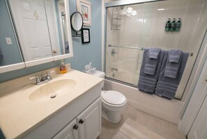 A bathroom with a sink, toilet, and shower. There are towels hanging on the wall and bottles of shampoo and conditioner.