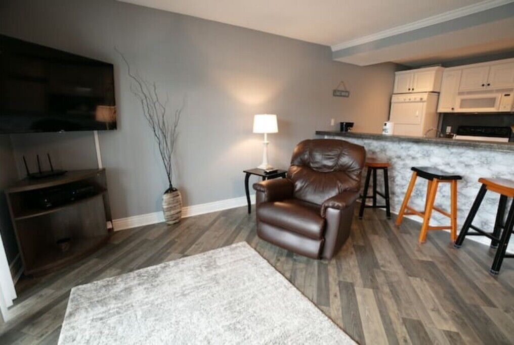 A living room with a leather chair, a TV, a lamp, and a kitchen area with stools.