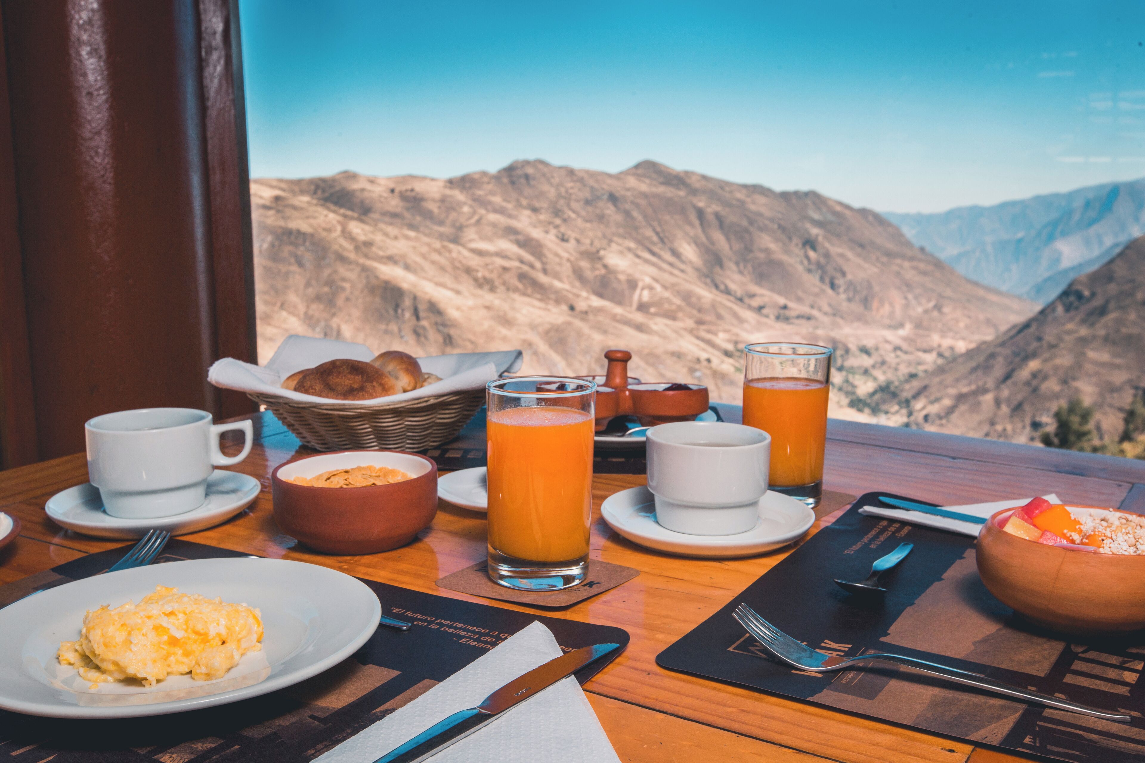A breakfast table with a mountain view, including a plate of scrambled eggs, a glass of orange juice, and a bowl of cereal.