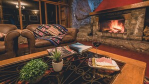 A cozy living room with leather chairs, a fireplace, a glass table with a plant, a book, and a plate of cheese and olives. - Refugio Viñak (Vinac)