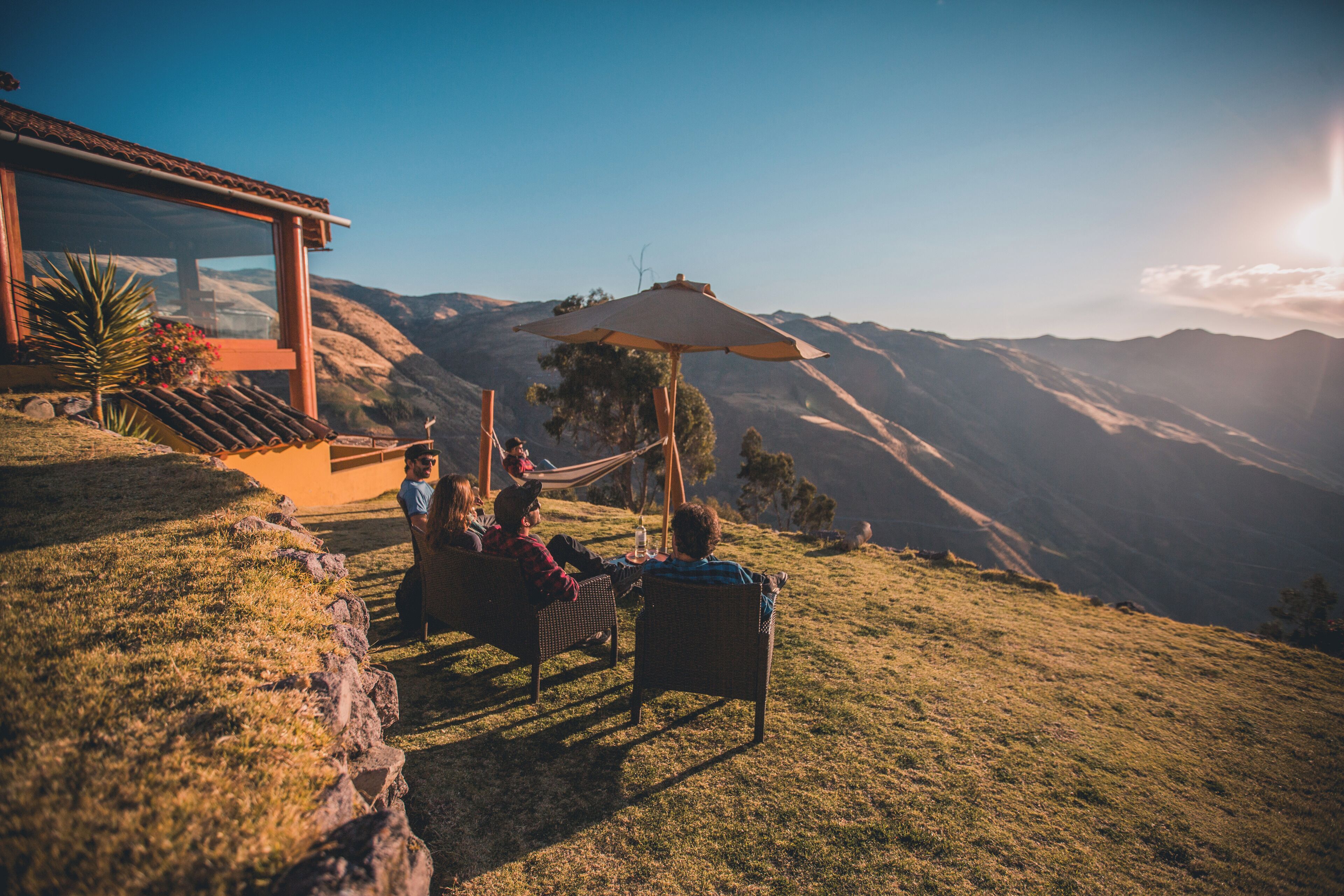 Un couple se détend dans un hamac, avec une vue sur les montagnes, devant un bâtiment en verre et sur une terrasse en bois.