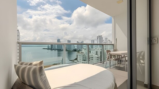 A balcony with a bed, a table, and chairs, overlooking a cityscape and water. - 16th Floor Infinitum 1 Bedroom Apt (Cartagena)