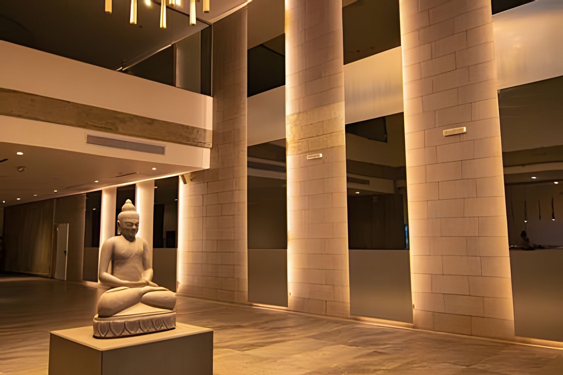 A modern interior with a large Buddha statue on a pedestal, surrounded by high ceilings and columns.