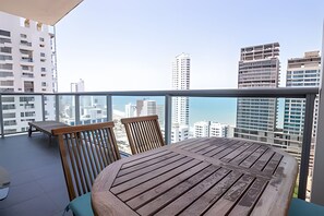 A balcony with a wooden table and chairs, overlooking a cityscape with tall buildings and a clear sky.