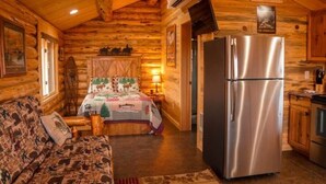 A log cabin interior with a wooden ceiling, a bed with a forest-themed bedspread, a sofa with animal prints, a stainless steel refrigerator, and a kitchen area with wooden cabinets. - The Bighole Cabin at J&J Cabins (Salmon)