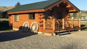 A log cabin with a porch, two large wooden wheels, and a gravel driveway. - The Bighole Cabin at J&J Cabins (Salmon)