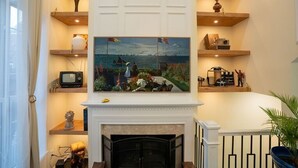 A living room with a fireplace, wooden shelves with decorative items, a painting above the fireplace, and a television on a shelf. - Luxury Townhome Minutes From DC (Alexandria)