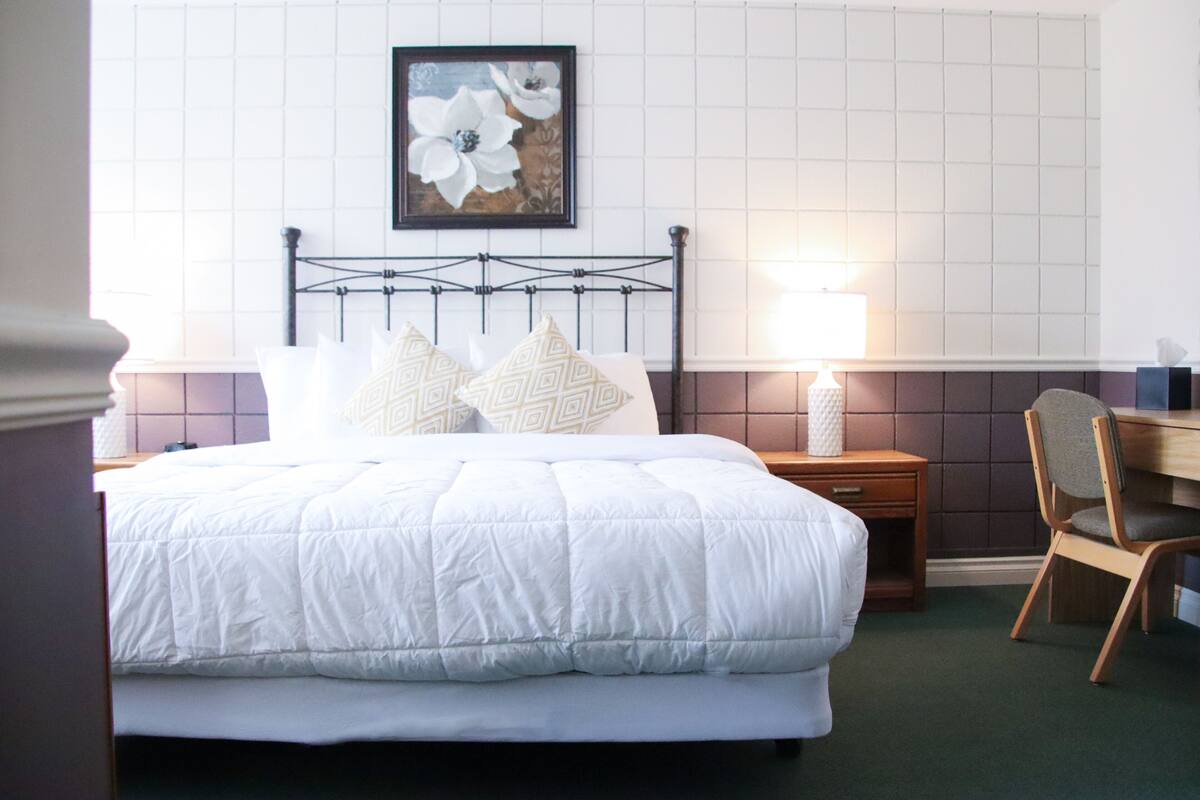 A neatly made bed with a white comforter and patterned pillows, a wooden nightstand with a lamp, and a chair with a cushion.