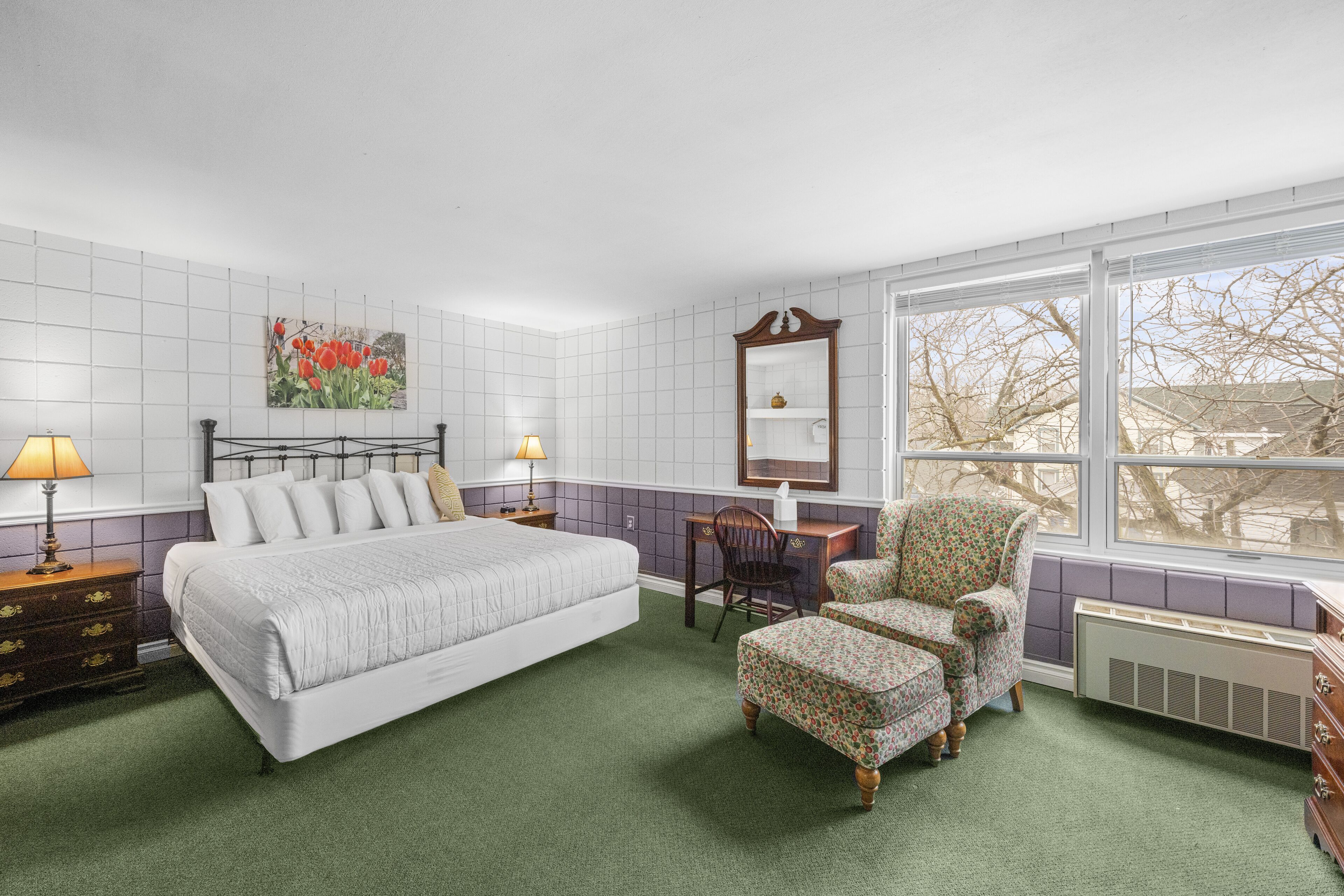 A bedroom with a large bed, two bedside tables with lamps, a chair, a dresser, a mirror, and a window with a view of trees.