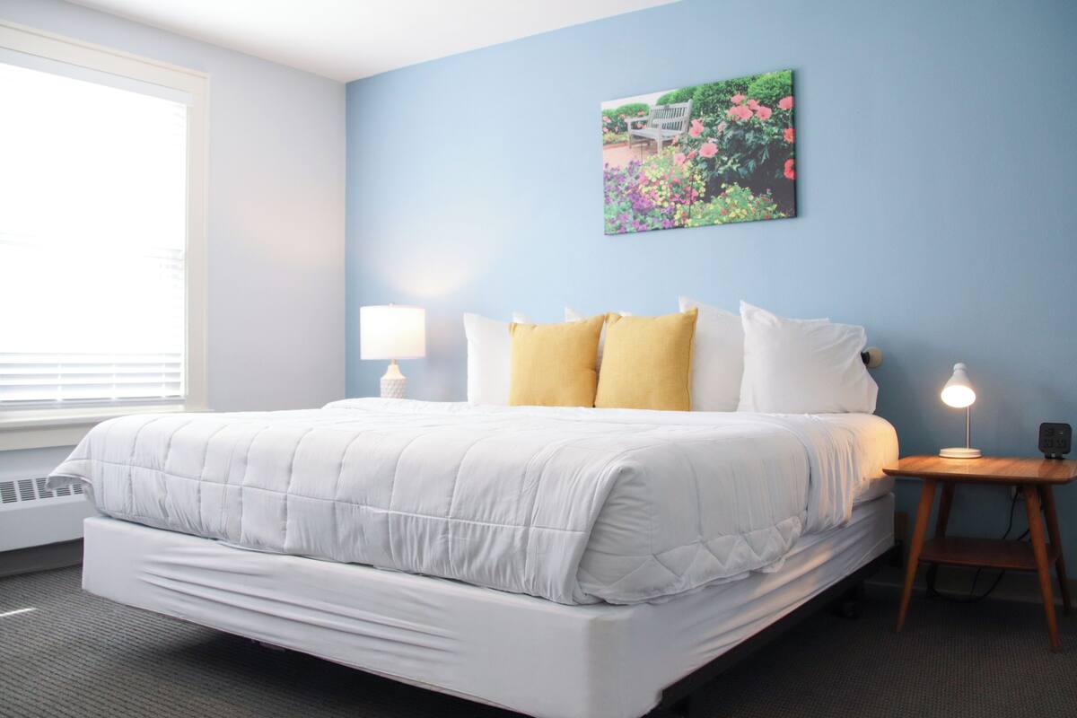 A neatly made bed with white and yellow pillows, a bedside lamp, and a wall-mounted picture of flowers.