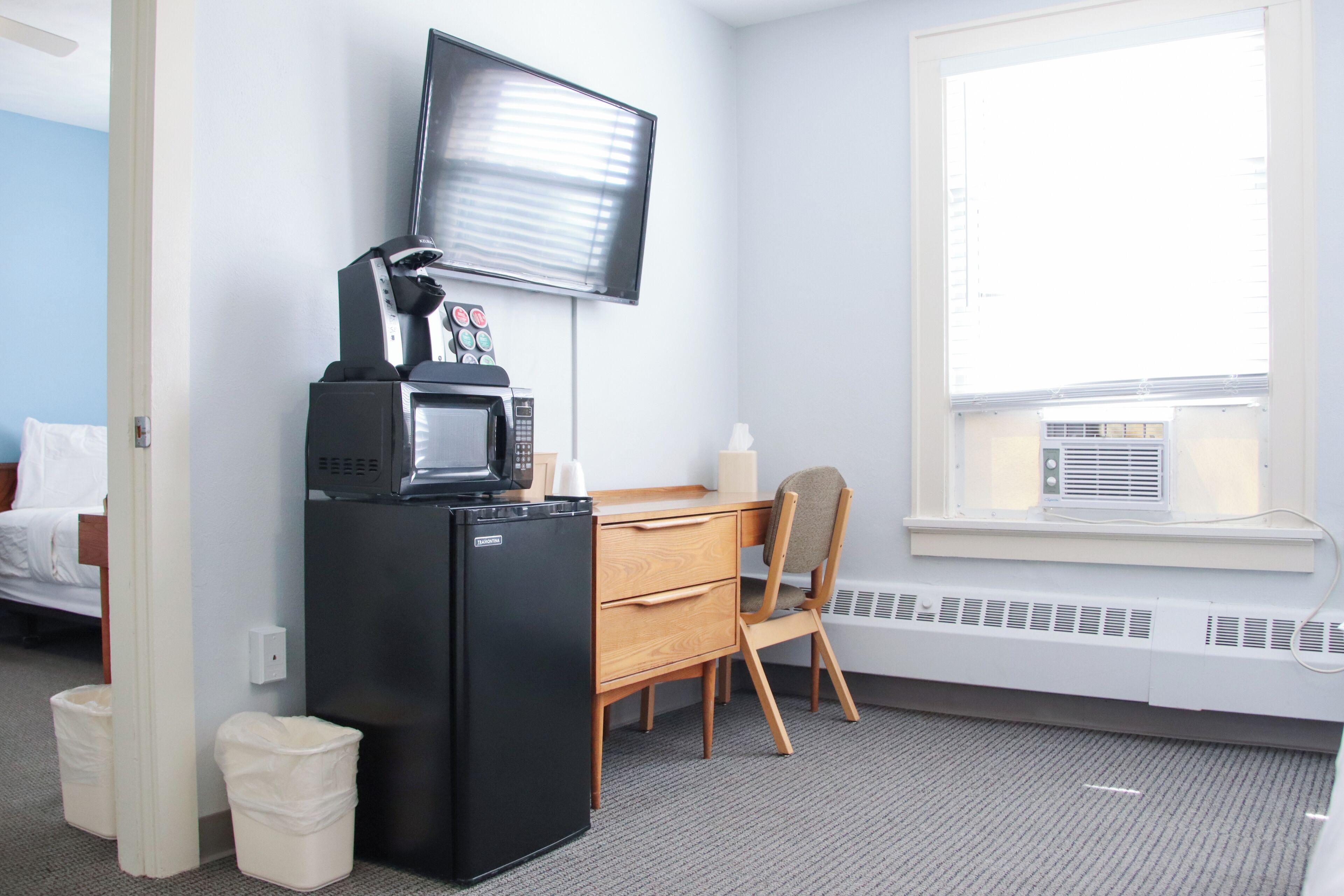 A hotel room with a kitchenette, a flat-screen TV mounted on the wall, a window with blinds, a desk with a chair, and a radiator under the window.
