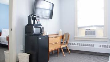 A hotel room with a kitchenette, a flat-screen TV mounted on the wall, a window with blinds, a desk with a chair, and a radiator under the window.
