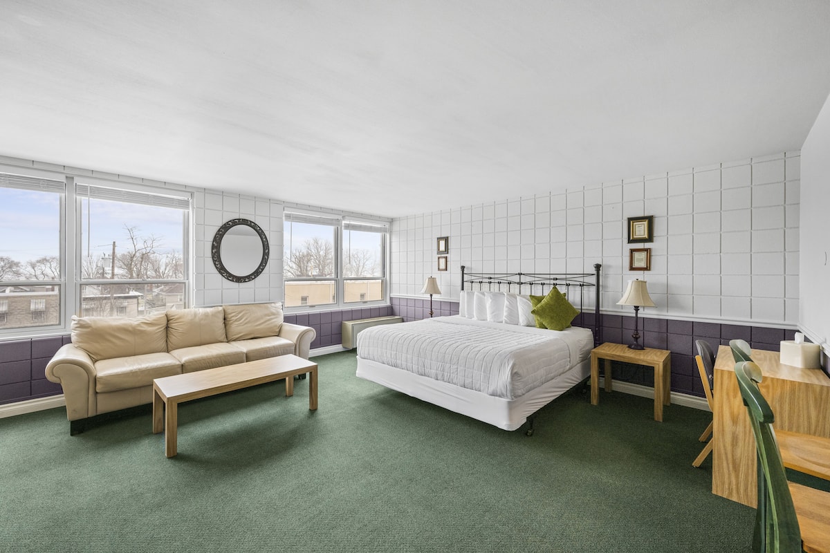 A modern hotel room with a large bed, a sofa, a small table, and a chair. There are windows providing a view of the outside.