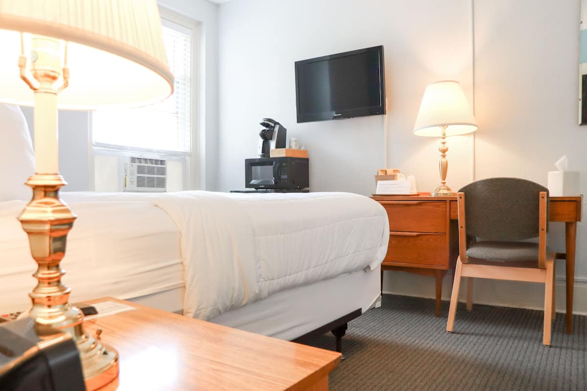 A hotel room with a bed, a desk, a chair, a TV, and a lamp.