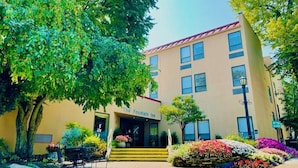 A hotel with a yellow facade, a red roof, and a sign reading "The Fountain Inn." There are steps leading to the entrance, surrounded by colorful flower beds and trees. - The Fountain Inn (Marblehead)