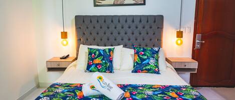 A neatly made bed with a tropical-themed bedspread, two matching pillows, a folded towel, and a framed artwork above the headboard.