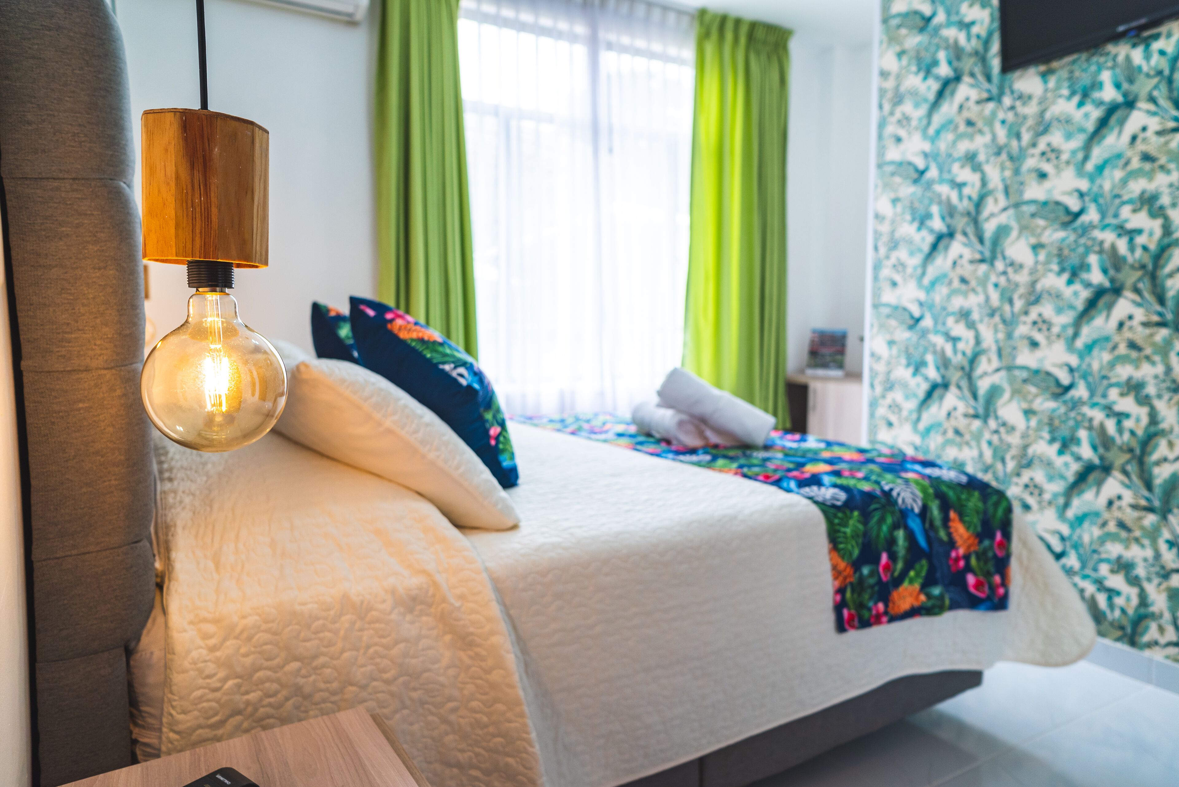 A neatly made bed with a white comforter and colorful pillows, a wooden bedside table, and a hanging light fixture.