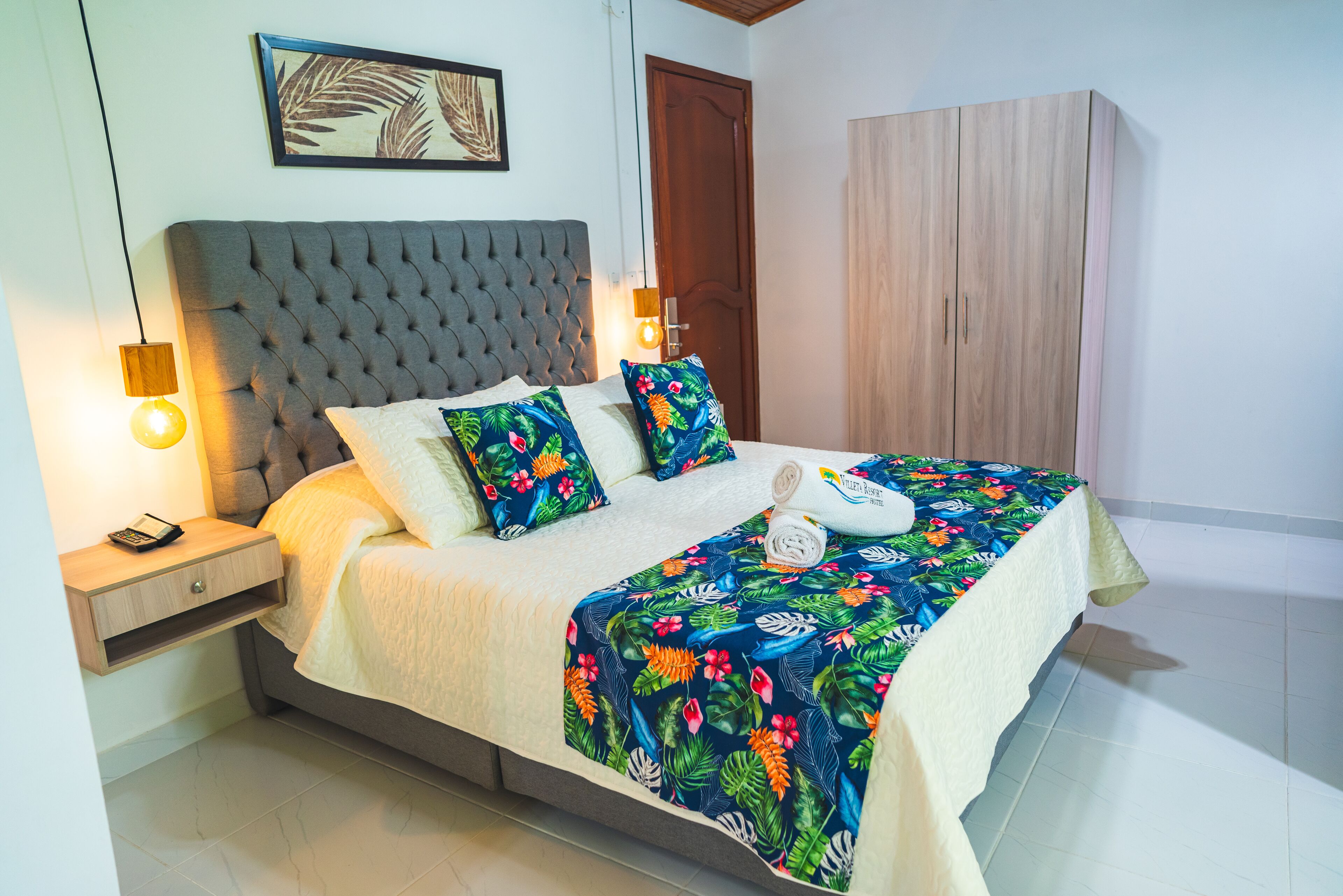 A neatly made bed with a floral bedspread, a bedside table with a lamp, and a wooden wardrobe.