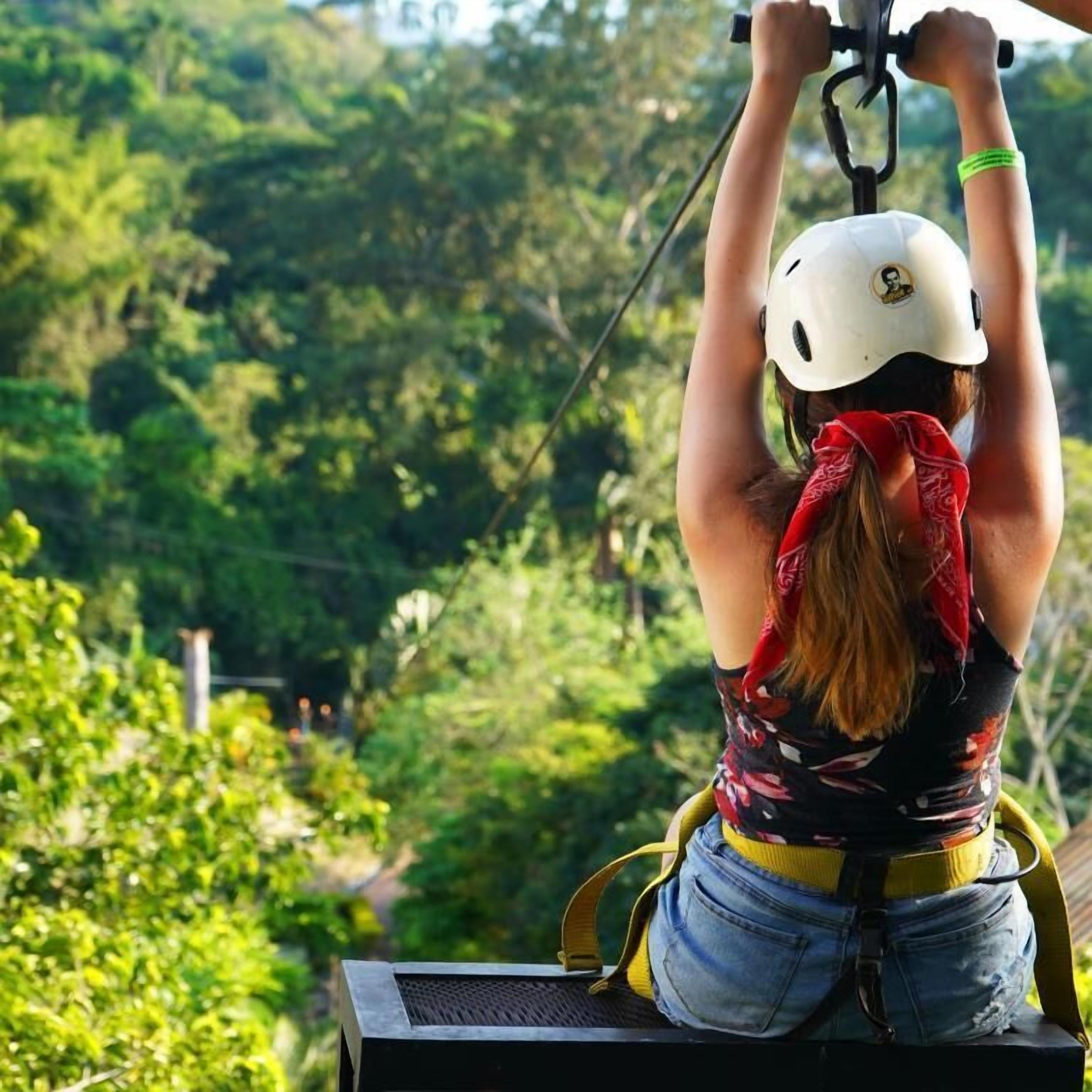 A person on a zip line with a helmet, harness, and red bandana, overlooking a forested area.