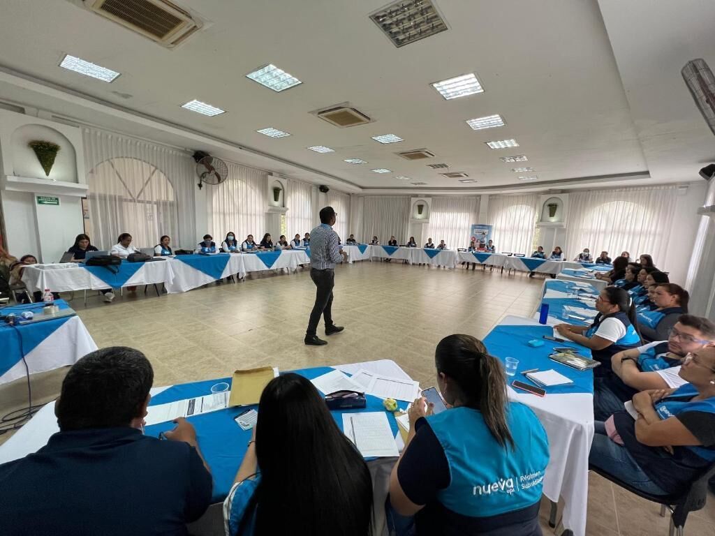 A conference room with round tables, attendees in blue vests, and a speaker at the front.