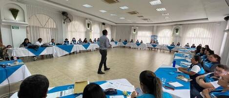 A conference room with round tables, attendees in blue vests, and a speaker at the front.