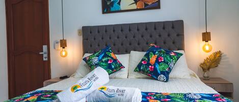 Una cama bien hecha con una colcha estampada con motivos florales, dos almohadas con estampados tropicales y un cuadro en la pared.